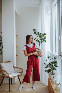 Young woman stands at home with a house plant in a pot in her hands