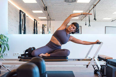 Slim adult female doing seated side bend and stretching body on modern reformer during pilates training in gym