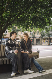 Happy male and female teenage friends laughing while sitting on bench under tree