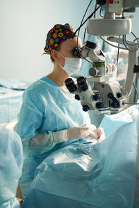 Adult female doctor in sterile mask and ornamental medical cap looking through surgical microscope against crop coworker in hospital