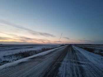 Road amidst snow covered landscape against sky during sunset