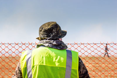 Rear view of soldier wearing uniform while standing by fence against sky