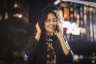 Happy young woman looking away while answering smart phone in city at night