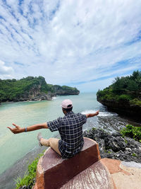 Rear view of a man sitting on rock by sea against sky