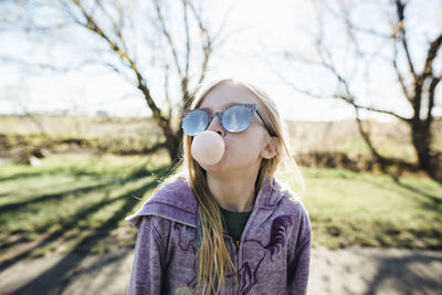 Girl wearing sunglasses blowing bubble gum while standing on road against sky