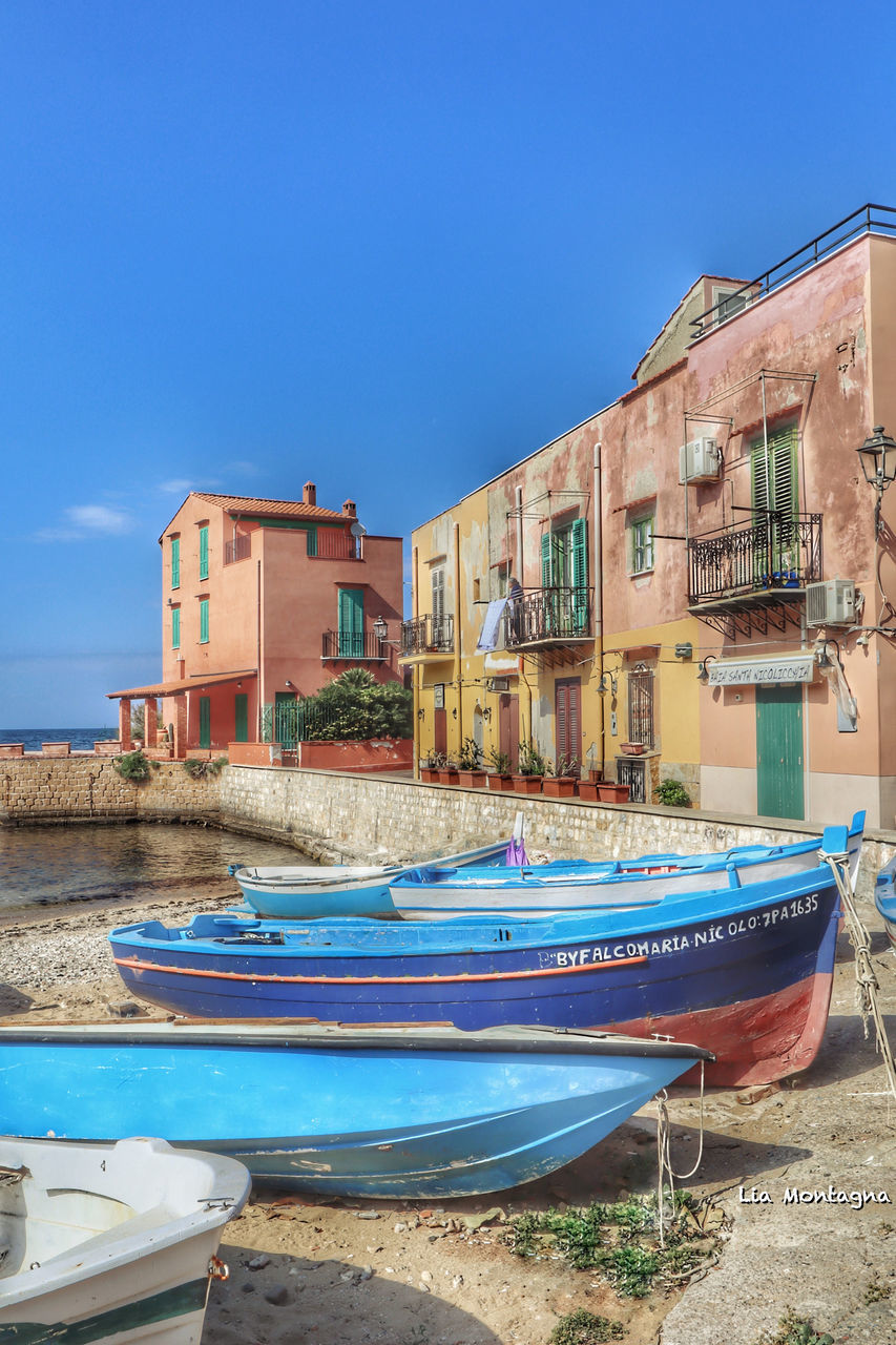 BOATS MOORED AT BEACH BY BUILDINGS AGAINST BLUE SKY