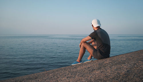A young man sits on a granite seashore. the sea is calm. the man looks to the horizon.