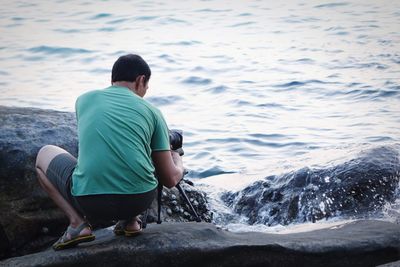 Rear view of man crouching while photographing on rocky shore