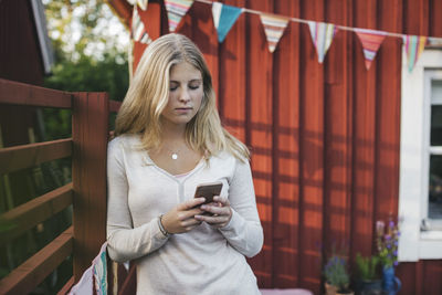 Teenage girl leaning on fence while text messaging against house in back yard