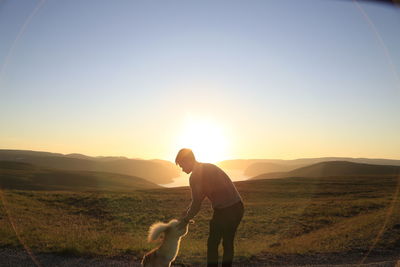 Rear view of man with dog on field against sky during sunset