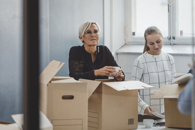 Mature businesswoman holding coffee cup while unpacking cardboard boxes in new office
