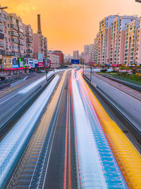 Light trails on city street by buildings against sky during sunset