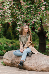 Cute teenage girl with long brown hair in raincoat is sitting on a rock in the park