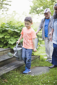 Boy carrying watering can with grandparents walking in back yard