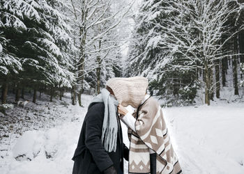 Romantic couple covering face with scarf while standing on snow in forest