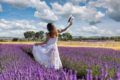 Woman seen from behind plays with her long white dress in the middle of a blooming lavender field.