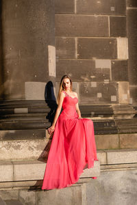 Full length of young woman in pink evening gown sitting against columns