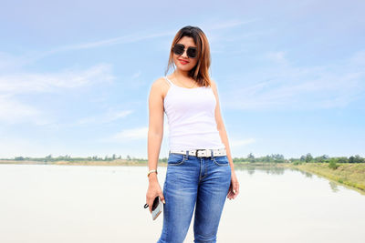 Portrait of young woman wearing sunglasses standing by lake against sky