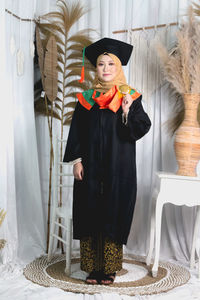 Portrait of smiling young woman wearing graduation gown standing against wall