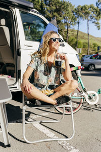 Cheerful woman enjoying a juice while sitting out of caravan in camping near surf board and bicycle