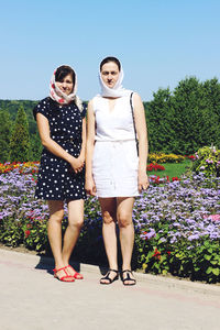 Full length portrait of happy friends standing by flowering plants clear sky