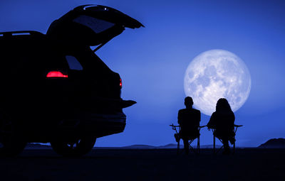 Couple seating next to their car and enjoying full moon vista during scenic desert night. 