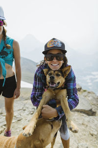 Portrait of woman embracing dog while standing by friend on mountain against clear sky