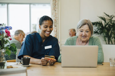 Smiling female caregiver assisting senior woman doing online shopping with credit card and laptop in nursing home