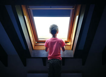 Rear view of boy looking through window while standing at home