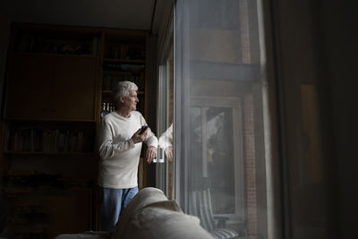 Senior man with mobile phone looking away while standing by window at home