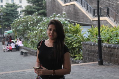 Portrait of young woman with mobile phone standing on footpath in city