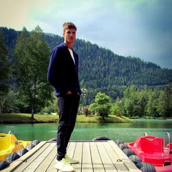 Portrait of young man standing by lake against trees