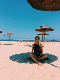 Portrait of woman sitting on sand at beach against clear blue sky