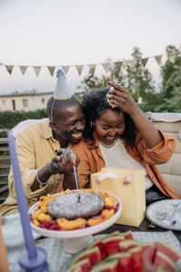 Happy young man sitting with female friend cutting cake while celebrating birthday in back yard