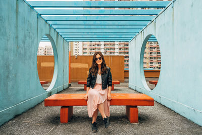 Portrait of young woman sitting on bench amidst wall