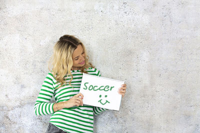 Smiling beautiful woman holding book with text while standing against wall