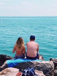 Rear view of man and woman sitting on rock while looking at sea against sky