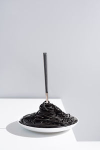 Stainless fork in full bowl of black spaghetti with cuttlefish ink on white table in studio on gray background