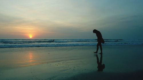 Silhouette man walking on shore against cloudy sky during sunset