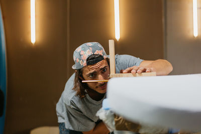 Concentrated male hipster using scribe tool with pencil while marking surfboard before shaping in workshop