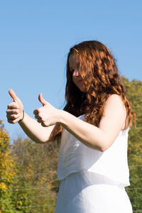 Low angle view of young woman showing thumbs up while standing against clear blue sky during sunny day