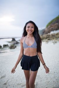 Portrait of smiling young woman standing at beach against sky