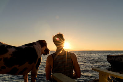 Rear view of man standing with dog against sea during sunset