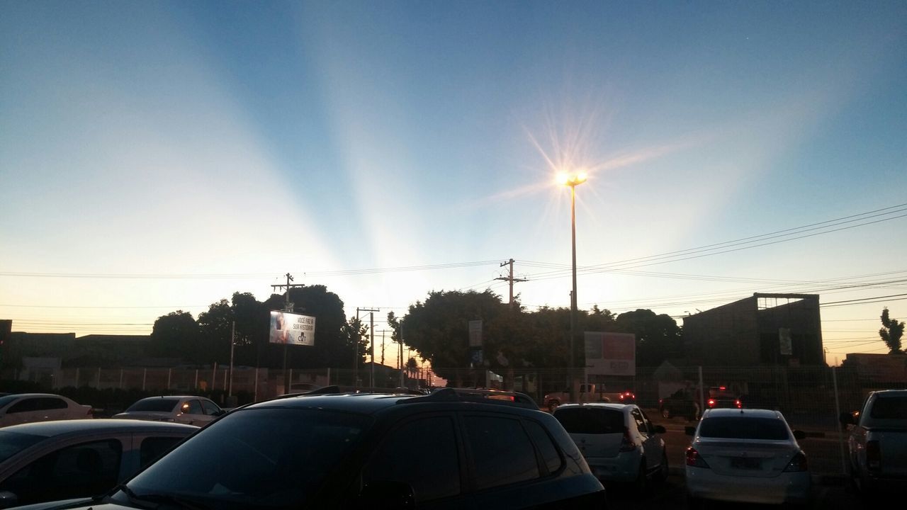 transportation, car, land vehicle, mode of transport, built structure, sky, building exterior, architecture, road, street, street light, sun, city, sunlight, sunbeam, cloud - sky, sunset, electricity pylon, lens flare, blue