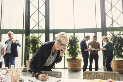 Mature businesswoman on call while writing in notepad with colleagues in background at office