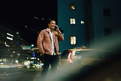 Smiling man talking through smart phone while standing in illuminated city at night