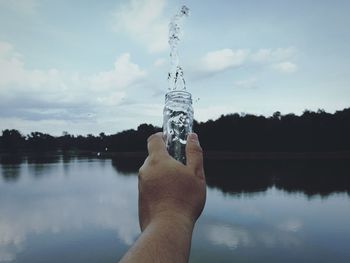 Cropped hand splashing water from bottle in lake against sky