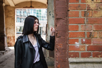 Sad young girl in black jacket and jeans stands in old arch on city street on summer day