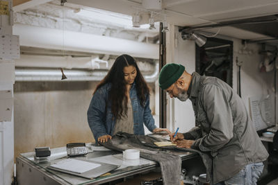 Male tailor writing bill while standing with customer at workbench