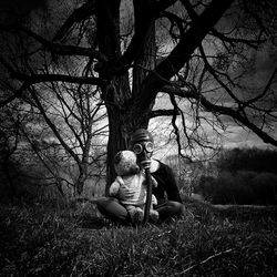 Teenage boy wearing gas mask holding stuffed toy sitting against tree trunk